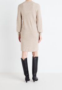 Beige ribbstickad klänning med långa ärmar, knälång, kombinerad med svarta knähöga stövlar med spetsig tå och V-cut-detalj.