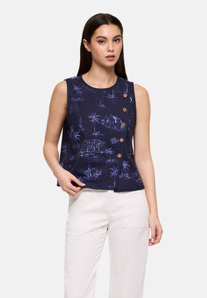 Mujer con camiseta sin mangas azul marino con botones de madera y estampado costero, combinada con pantalones blancos, posando frente a un fondo claro.