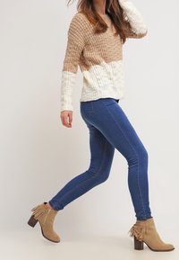 Suéter de punto con diseño ombré en tonos beige y marrón, combinado con jeans ajustados azules y botines color tan con flecos y tacón cuadrado.