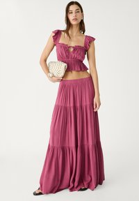Zweiteiliges Outfit in staubigem Rosé. Oberteil mit Rüschensäumen und Schlüssel-Ausschnitt; gestufter Maxirock mit elastischem Taillenbund. Hält eine Perlenhandtasche.