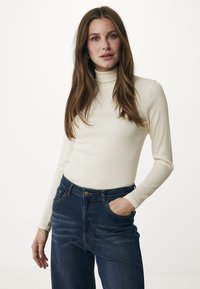 Cremfarbener gerippter Rollkragenpullover kombiniert mit dunkelblauen hochtaillierten Jeans. Der Pullover hat lange Ärmel und eine körpernahe Silhouette.