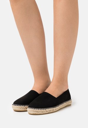 Espadrilles en suède noir avec un design à encolure basse, semelle en corde de jute et détails cousus le long du bord, posées sur des jambes nues.