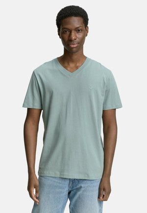 Camiseta de pico en verde menta, hecha de un tejido suave, con mangas cortas y un pequeño logo en el pecho, combinada con unos vaqueros azul claro.