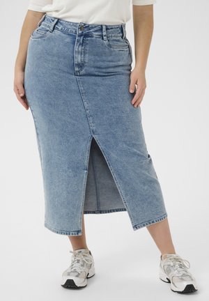 Kvinde iført lyseblå denim midi-nederdel med frontslids, hvid top og hvide sneakers mod ensfarvet baggrund.