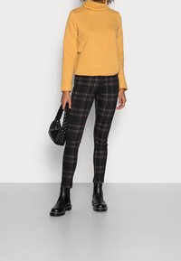 jersey de cuello alto amarillo, pantalones slim-fit de cuadros negros con detalles en rojo y azul, botas de tobillo de cuero negro y un bolso trenzado negro.