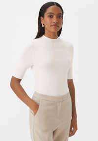 Haut blanc à manches courtes avec un col montant, en tissu lisse, associé à un pantalon beige clair avec des poches latérales.