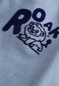 Tela azul claro con un gráfico de un león azul marino oscuro con una expresión sonriente, acompañado del texto "ROAR" en letras gruesas.