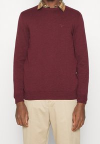 Pull bordeaux à col ras du cou et poignets côtelés, porté par-dessus une chemise à carreaux aux motifs beige, noir et blanc.
