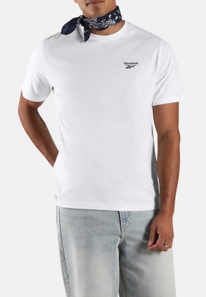 Man draagt een wit Reebok T-shirt, lichtblauwe jeans, een zwarte ring aan zijn rechterhand en een donkergepatteerd bandana om de nek geknoopt.