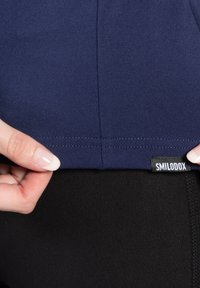 Marineblauw sporttopje met een gladde textuur en een gebogen zoom, voorzien van een zichtbaar merkembleem. Draagt over zwarte legging.