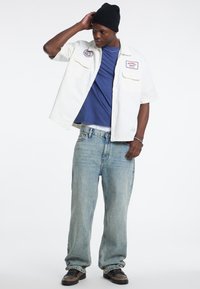 Camicia bianca a maniche corte sopra una maglietta blu, abbinata a jeans azzurri chiari e scarpe da ginnastica scure, con toppe e un fit rilassato.