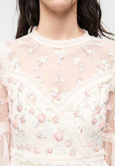 Κοντινό πλάνο ενός διαφανούς λευκού δαντελωτού τοπ με κεντήματα λουλουδιών σε απαλό ροζ και πράσινο, με βολάν γύρω από το ντεκολτέ και τα μανίκια.