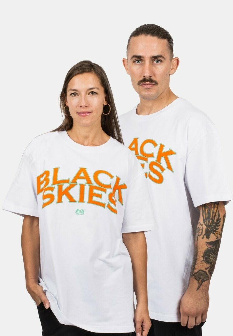 Hvide bomulds t-shirts med fed orange tekst "BLACK SKIES" og et grønt skald grafik. Løs pasform med korte ærmer og rund hals.