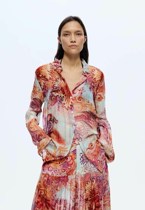 Vrouw draagt een kleurrijke blouse met lange mouwen en knopen en een bijpassende rok met abstracte bloemen- en wervelpatronen, handen in de zakken.
