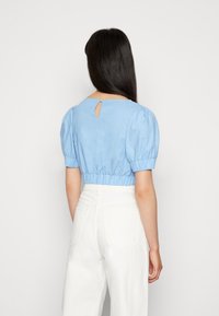 Blusa cropped azul claro com mangas bufantes, barra elástica e um pequeno detalhe em forma de fenda nas costas. Combinada com calças brancas de cintura alta.