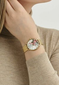 Goldfarbene Armbanduhr mit floralem Design auf einem weißen Zifferblatt, ausgestattet mit einem Metallarmband und einem glatten runden Gehäuse. Strukturierter Strickpullover im Hintergrund.