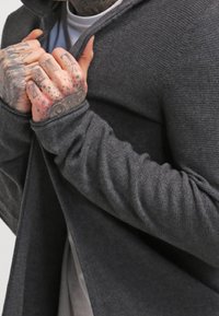 Tmavě šedý pletený cardigan s kapucí, s ribrovou texturou, otevřeným předním dílem a otvory pro palce na manžetách. Detailní záběr na ruce svírající tkaninu.