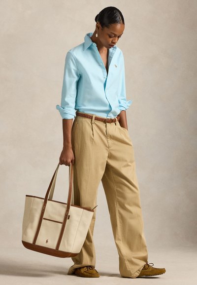 Frau mit hellblauem Hemd und beigen plissierten Hosen, hält eine beige-braune Tragetasche und steht vor neutralem Hintergrund.