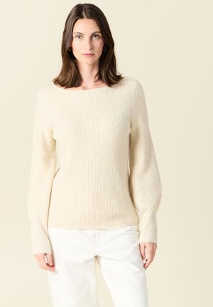 Beiger, gerippter Pullover mit Boot-Ausschnitt und langen Ärmeln. Aus weichem, strukturiertem Stoff gefertigt. Kombiniert mit weißen Hosen.