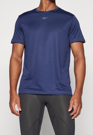 Uomo che indossa una maglietta sportiva aderente blu navy e leggings compressivi grigio scuro, in piedi davanti a uno sfondo chiaro e uniforme.