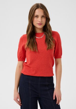 Femme aux longs cheveux ondulés portant un haut rouge texturé à manches courtes, un pantalon sombre taille haute et un collier argenté, se tenant devant un fond uni.