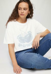 Biała koszulka z bawełny z jasnoniebieską grafiką wiśni i napisem "posiadaj swoją moc". Model w niebieskich dżinsach, siedzący.