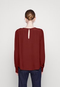 Bordeauxrot-blus mit langen Ärmeln, Schlüssel-Ausschnitt auf der Rückseite, rundem Halsausschnitt und leicht gerafften Ärmeln, aus einem glatten, leichten Stoff gefertigt.