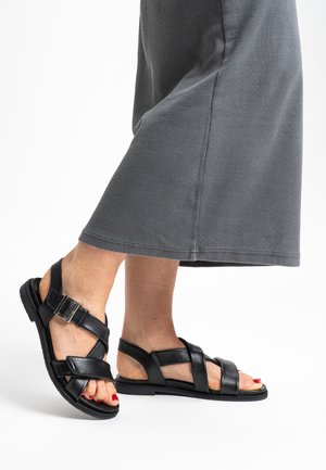 Feet wearing black leather sandals with buckles, paired with gray ribbed wide-leg pants against a white background.