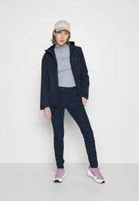 Giacca nera impermeabile con cappuccio regolabile sopra una felpa grigia chiara con zip. Pantaloni scuri e scarpe sportive con accenti rosa. Design semplice.