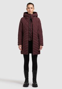 Abrigo acolchado burdeos con capucha, con un patrón diagonal, cierre frontal de cremallera y bolsillos laterales. Lleva puesto sobre leggings negros y botas.