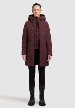 Bordeaux gewatteerde pufferjas met capuchon, voorzien van een diagonaal patroon, een ritssluiting aan de voorkant en zijzakken. Draagt over zwarte leggings en laarzen.