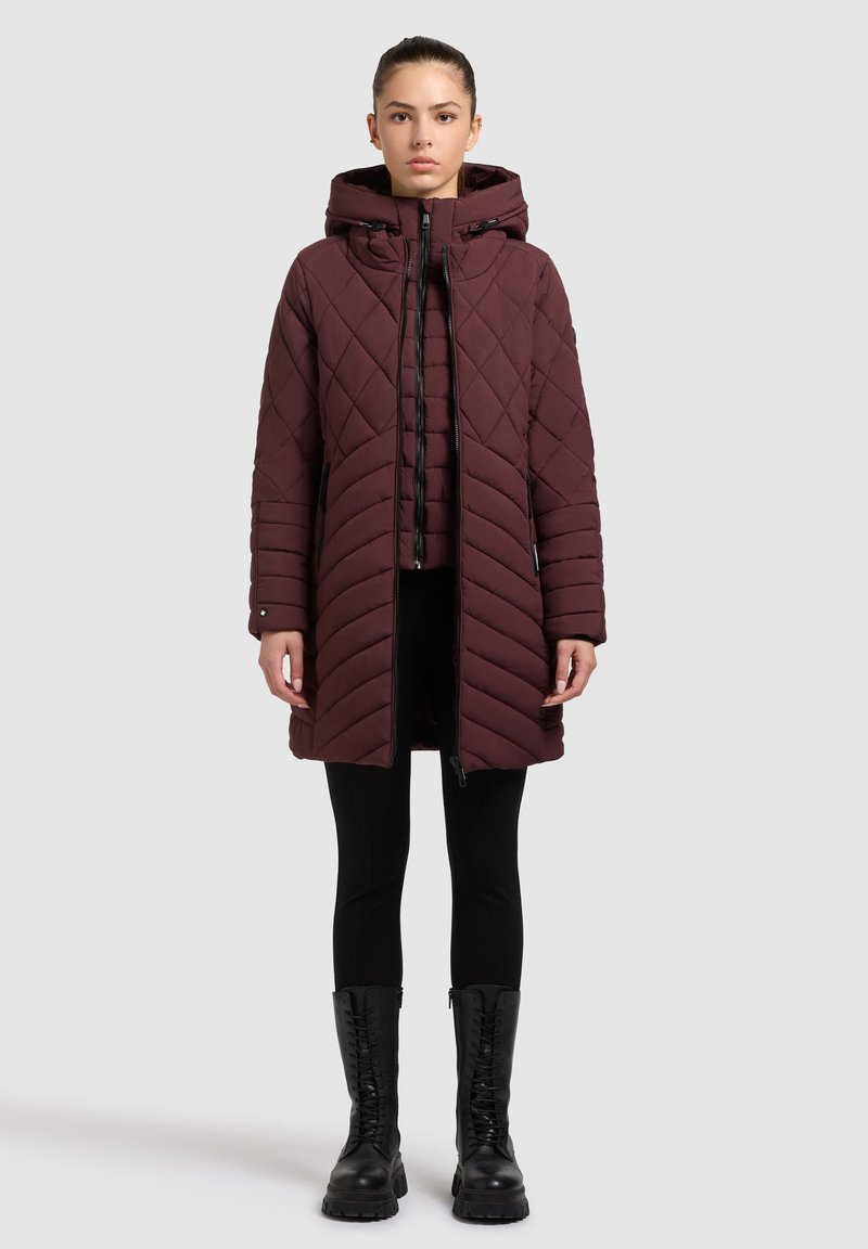 Abrigo acolchado burdeos con capucha, con un patrón diagonal, cierre frontal de cremallera y bolsillos laterales. Lleva puesto sobre leggings negros y botas.