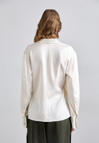 Blusa branca de manga longa com gola, textura suave, detalhe na costura das costas e punhos com botões, usada com calças verdes escuras.