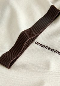 Cream-colored fabric with dark brown ribbed trim and "CREATIVE STUDIO" embroidered in dark brown thread.