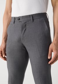 Homme portant un pantalon de costume gris slim avec un bouton et des passants de ceinture, associé à une chemise blanche rentrée.