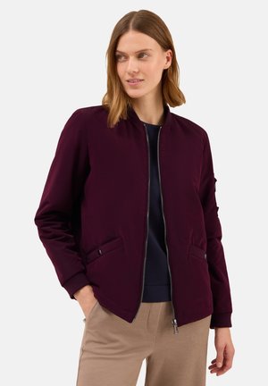 Frau trägt eine dunkelviolette Zip-Jacke über einem schwarzen Oberteil und beigen Hosen, mit Händen in den Taschen und schaut leicht zur Seite.