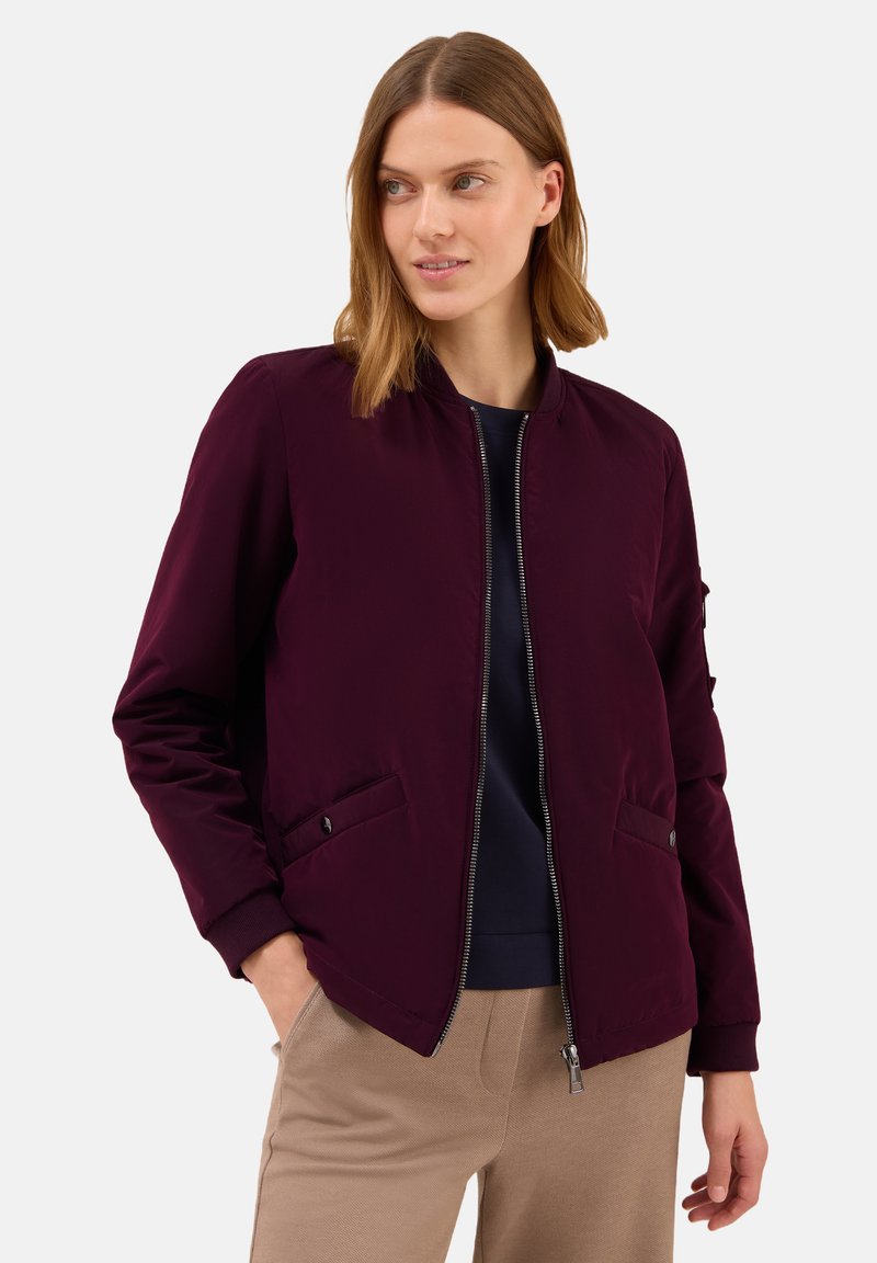 Woman wearing a dark purple zip-up bomber jacket over a black top and beige pants, with hands in pockets and looking slightly to the side.