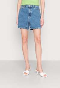 Persona con pantaloni di jeans blu, top verde e sandali bianchi con tacco, in piedi su un pavimento semplice.