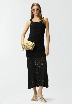 Femme portant une robe noire en dentelle sans manches, des sandales beige à talons hauts, de grandes lunettes de soleil et tenant un petit sac à main jaune.