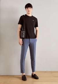 Svart t-shirt, blå skräddarsydda byxor och svarta loafers. En läderväska bärs över axeln. Modellen står framför en ljusgrå vägg.