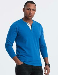 Man med kort hår som bär en blå långärmad henley-tröja och svarta jeans, stående med ena handen i fickan mot vit bakgrund.