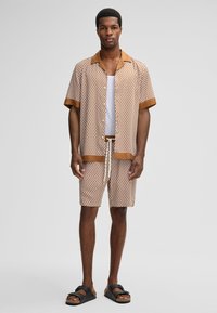 Uomo in camicia a maniche corte a fantasia marrone e pantaloni corti coordinati, canotta bianca e sandali neri con fibbia in piedi contro uno sfondo semplice.