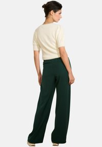 Kremo spalvos trumpomis rankovėmis megztinis su tekstūruotu paviršiumi, derinamas su tamsiai žaliais, platėjančiais kelnais su elastine juosmens juosta.