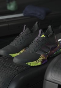 Paire de chaussures de sport noires avec semelles à motifs vert néon et violet placées sur un tapis roulant dans une salle de sport.