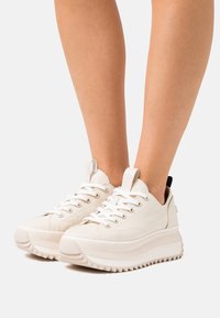 Bílé plátěné tenisky s tlustou podrážkou, s texturovaným vzorem, zaoblenou špičkou a kontrastním černým patním páskem. Klasické šněrování, nízký střih.