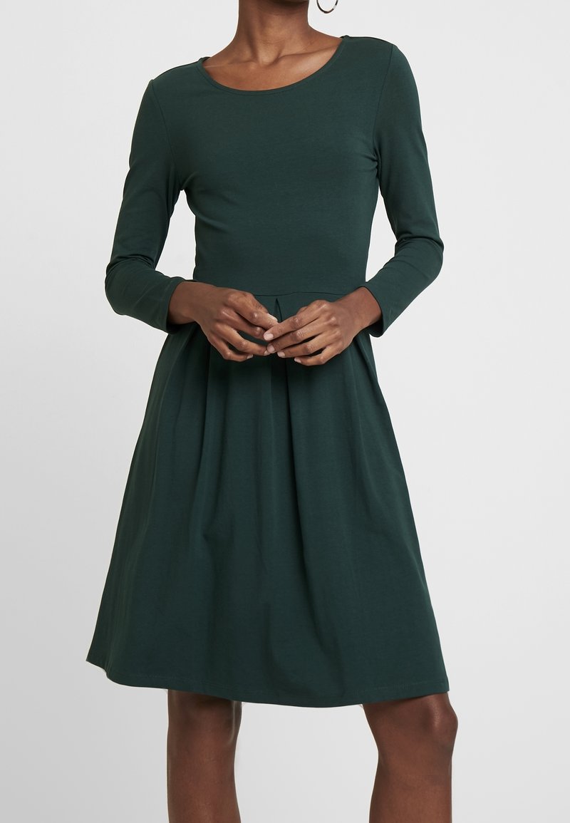 Femme portant une robe vert foncé à manches longues avec un corsage ajusté et une jupe plissée en ligne A, mains jointes devant elle.