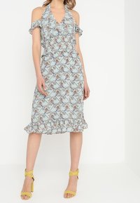 Ljusblå blommig klänning med cold-shoulder design, volangärmar, figurnära midja och fransad nederkant. Matchas med gula sandaler.