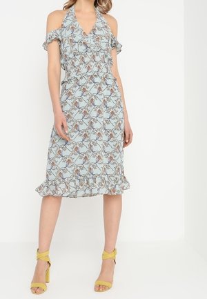 Robe florale bleu clair avec un design à épaules dénudées, des manches volantées, une taille cintrée et un ourlet frangé. Associée à des sandales jaunes.