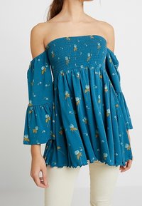 Turkoosi olkapäät paljastava yläosa, kevyt kangas, jossa on kerrostettua rypytystä, kellohihat ja valkoisilla ja keltaisilla korostuksilla koristeltu kukkakuvio.