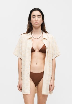 Žena v hnedom bikine s béžovou vzorovanou krátkou košeľou otvorenou a bielym korálkovým náhrdelníkom, stojaca pred jednoliatym pozadím.